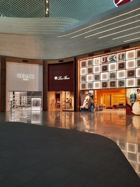 A shopping mall interior featuring high-end luxury fashion stores. The storefronts include Celine, Loro Piana, and Gucci. The design is modern with an illuminated ceiling and glossy floors reflecting the ambient lighting. Display windows showcase fashion items, and the space appears spacious and well-organized.