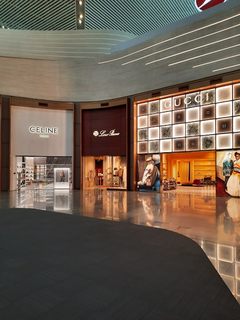 A shopping mall interior featuring high-end luxury fashion stores. The storefronts include Celine, Loro Piana, and Gucci. The design is modern with an illuminated ceiling and glossy floors reflecting the ambient lighting. Display windows showcase fashion items, and the space appears spacious and well-organized.