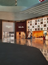 A shopping mall interior featuring high-end luxury fashion stores. The storefronts include Celine, Loro Piana, and Gucci. The design is modern with an illuminated ceiling and glossy floors reflecting the ambient lighting. Display windows showcase fashion items, and the space appears spacious and well-organized.