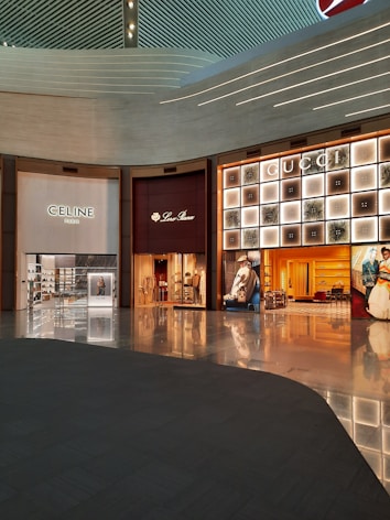 A shopping mall interior featuring high-end luxury fashion stores. The storefronts include Celine, Loro Piana, and Gucci. The design is modern with an illuminated ceiling and glossy floors reflecting the ambient lighting. Display windows showcase fashion items, and the space appears spacious and well-organized.