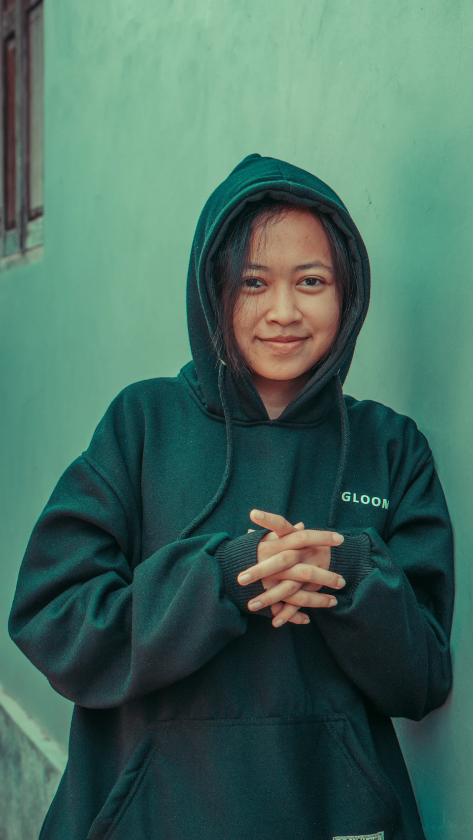 Perempuan berhoodie hijau tersenyum di depan dinding biru kehijauan