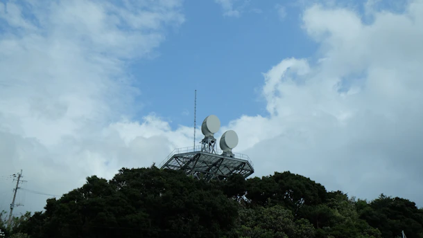 A metal structure with two large satellite dishes is situated on top of a hill covered with dense trees. The sky above is a mix of blue and white clouds, suggesting a partly cloudy or clearing weather.