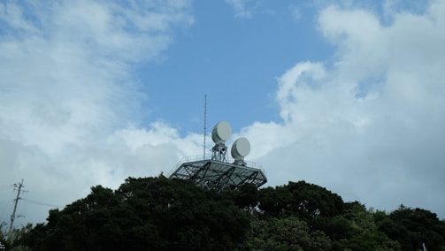 A metal structure with two large satellite dishes is situated on top of a hill covered with dense trees. The sky above is a mix of blue and white clouds, suggesting a partly cloudy or clearing weather.