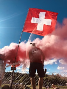 A person stands behind a fence holding a large Swiss flag, with red smoke enveloping the area. The sky is clear and blue, and there are some trees visible in the distance. The person is wearing a shirt with '91' and 'Seewer' printed on the back, and is surrounded by vibrant red and white colors.