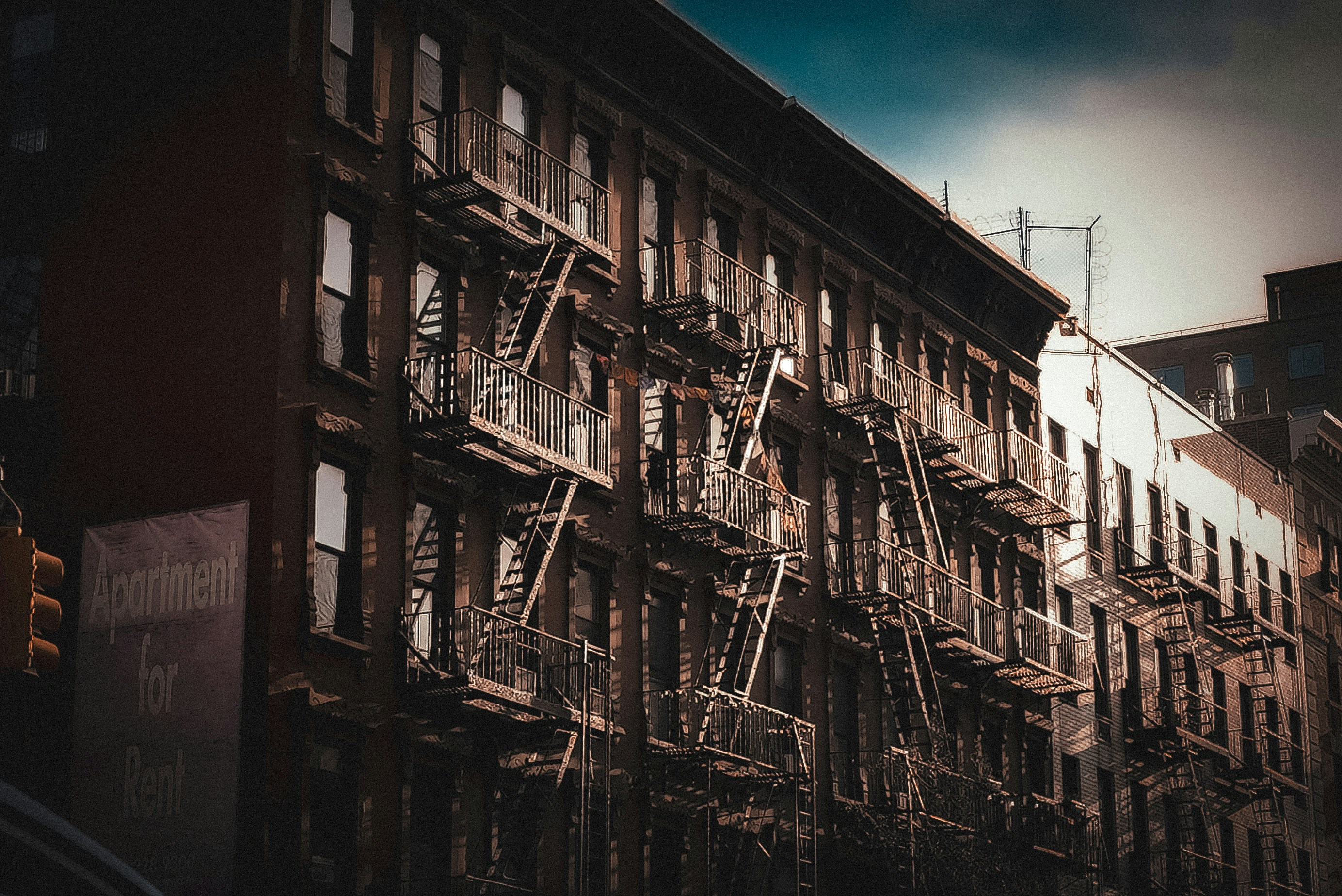 Sunlit apartment building with fire escapes, featuring a "For Rent" sign; the facade displays a mix of shadows and light.