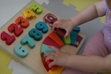 A child is playing with a colorful wooden puzzle board featuring numbers and geometric shapes. The numbers range from 1 to 9 in various bright colors like red, orange, yellow, and green. Next to the numbers, there are rainbow-colored semicircles arranged on a wooden base. The child's hands are interacting with the puzzle pieces, and the child is wearing a purple outfit with white polka dots on a textured play mat.