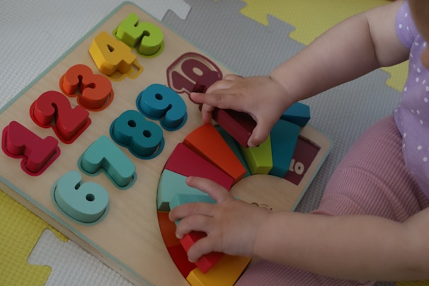 A child is playing with a colorful wooden puzzle board featuring numbers and geometric shapes. The numbers range from 1 to 9 in various bright colors like red, orange, yellow, and green. Next to the numbers, there are rainbow-colored semicircles arranged on a wooden base. The child's hands are interacting with the puzzle pieces, and the child is wearing a purple outfit with white polka dots on a textured play mat.