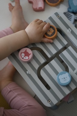 A child's hand is interacting with a wooden toy puzzle featuring colorful animal faces. The toy has striped patterns and pieces shaped like a lion, a fox, and a bear. The child is wearing pink pants, and the background shows multiple other toy pieces scattered around.
