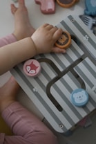 A child's hand is interacting with a wooden toy puzzle featuring colorful animal faces. The toy has striped patterns and pieces shaped like a lion, a fox, and a bear. The child is wearing pink pants, and the background shows multiple other toy pieces scattered around.