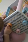A young child is engaging with a pastel-colored activity board featuring removable pieces with animal illustrations. The pieces are circular and designed to fit into specific slots. The child's clothing is soft purple, complementing the subtle tones of the board.