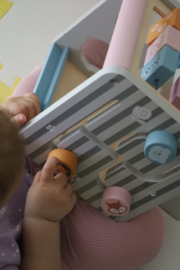 A young child is engaging with a pastel-colored activity board featuring removable pieces with animal illustrations. The pieces are circular and designed to fit into specific slots. The child's clothing is soft purple, complementing the subtle tones of the board.