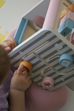 A young child is engaging with a pastel-colored activity board featuring removable pieces with animal illustrations. The pieces are circular and designed to fit into specific slots. The child's clothing is soft purple, complementing the subtle tones of the board.