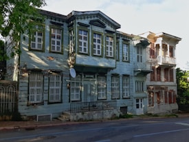 An old, weathered wooden house with peeling paint stands alongside a more well-maintained adjacent building. The facade features multiple windows with green shutters, and there is an overgrown garden partially visible. The architectural style is reminiscent of traditional wooden houses. The house appears deserted, and its aged look contrasts with the sunny environment.