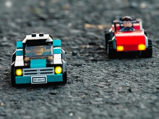 Two toy cars made of LEGO pieces are positioned on a textured surface resembling asphalt. The car in the foreground is blue with a LEGO figure inside, while the car in the background is red, also with a driver figure. Both cars have front lights that appear to be lit.