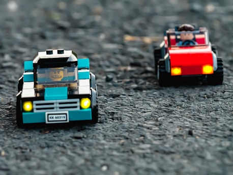 Two toy cars made of LEGO pieces are positioned on a textured surface resembling asphalt. The car in the foreground is blue with a LEGO figure inside, while the car in the background is red, also with a driver figure. Both cars have front lights that appear to be lit.