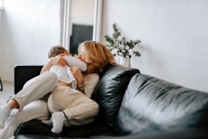 A cozy living room scene with a mother and child embracing on a black leather sofa. The room is softly lit, with a large mirror and potted plant in the background adding to the serene atmosphere. Both are wearing light-colored clothing, emphasizing the warmth and intimacy of the moment.