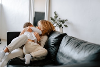 A cozy living room scene with a mother and child embracing on a black leather sofa. The room is softly lit, with a large mirror and potted plant in the background adding to the serene atmosphere. Both are wearing light-colored clothing, emphasizing the warmth and intimacy of the moment.