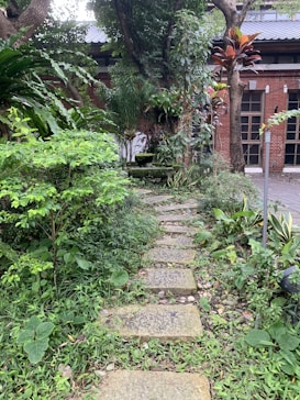 A serene garden path made of stone slabs leads through lush greenery.&nbsp;A variety of plants and trees surround the path, creating a peaceful, natural environment. A brick building is partially visible in the background, adding a rustic touch to the scene.