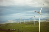 A landscape featuring a field with several wind turbines spread across the scene. The turbines are situated on a lush, green landscape with rolling hills in the background. The sky is overcast with clouds, giving a sense of a breezy day.