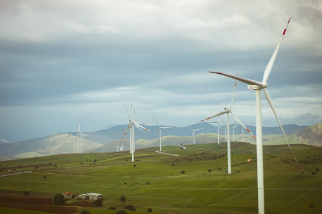 A landscape featuring a field with several wind turbines spread across the scene. The turbines are situated on a lush, green landscape with rolling hills in the background. The sky is overcast with clouds, giving a sense of a breezy day.