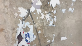 A surface with peeling paint showing multiple layers of colors including white, blue, and red. The texture is rough with visible patches and chips, revealing the underlying material.