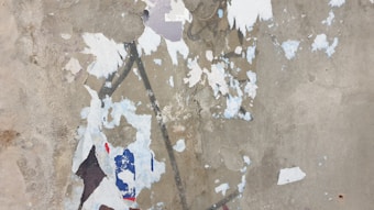 A surface with peeling paint showing multiple layers of colors including white, blue, and red. The texture is rough with visible patches and chips, revealing the underlying material.