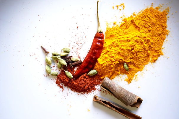 A collection of spices is artfully arranged on a white background. Visible are a red chili pepper, a pile of bright yellow turmeric powder, a smaller pile of orange-red chili powder, green cardamom pods, cloves, and cinnamon sticks.