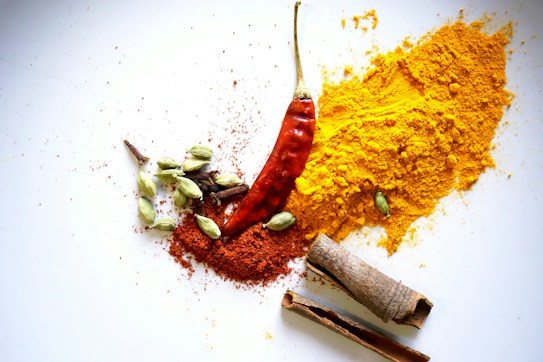 A collection of spices is artfully arranged on a white background. Visible are a red chili pepper, a pile of bright yellow turmeric powder, a smaller pile of orange-red chili powder, green cardamom pods, cloves, and cinnamon sticks.