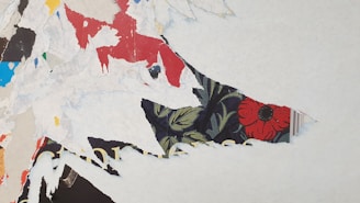 Torn layers of colorful paper create a textured collage with visible pieces of red, blue, and black paper. A prominent section includes a vibrant floral pattern featuring a red flower on a dark background, partially concealed by white, frayed paper edges.