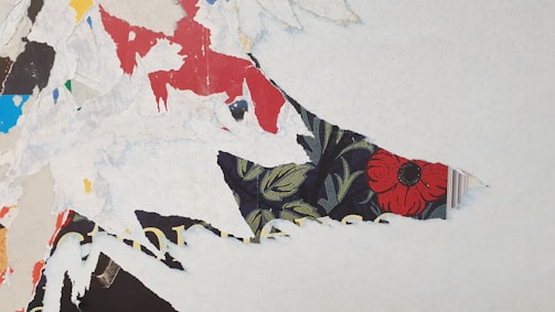Torn layers of colorful paper create a textured collage with visible pieces of red, blue, and black paper. A prominent section includes a vibrant floral pattern featuring a red flower on a dark background, partially concealed by white, frayed paper edges.
