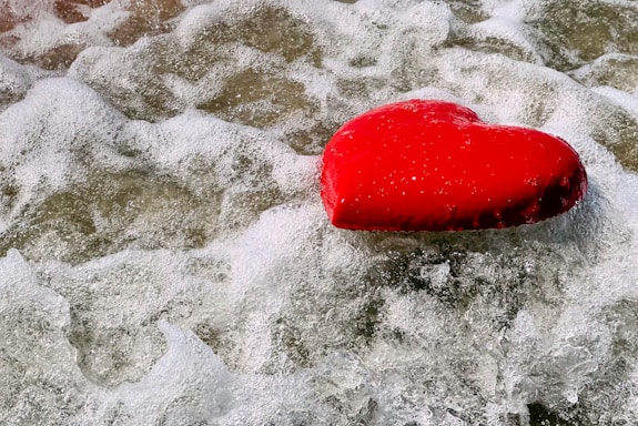 A bright red heart-shaped object is floating on foamy waves, surrounded by water in motion, creating a contrast between the vibrant color of the heart and the swirling white and gray patterns of the water.