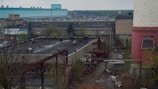 An industrial area featuring several large buildings, including a prominent structure with a teal roof and another with a pink and white exterior. The scene includes various industrial equipment, piping, and some trees. The environment appears to be somewhat overcast with muted lighting.
