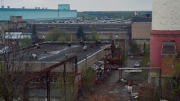 An industrial area featuring several large buildings, including a prominent structure with a teal roof and another with a pink and white exterior. The scene includes various industrial equipment, piping, and some trees. The environment appears to be somewhat overcast with muted lighting.