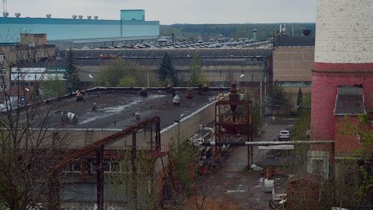 An industrial area featuring several large buildings, including a prominent structure with a teal roof and another with a pink and white exterior. The scene includes various industrial equipment, piping, and some trees. The environment appears to be somewhat overcast with muted lighting.