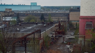 An industrial area featuring several large buildings, including a prominent structure with a teal roof and another with a pink and white exterior. The scene includes various industrial equipment, piping, and some trees. The environment appears to be somewhat overcast with muted lighting.