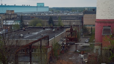 An industrial area featuring several large buildings, including a prominent structure with a teal roof and another with a pink and white exterior. The scene includes various industrial equipment, piping, and some trees. The environment appears to be somewhat overcast with muted lighting.
