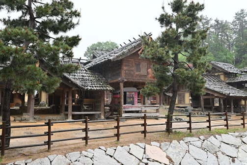 A traditional building with wooden architecture and a tiled roof, surrounded by several pine trees. The setting appears to be peaceful and rustic, with a natural stone wall in the foreground and a dirt pathway. The atmosphere suggests a historical or cultural context, possibly a village or heritage site.