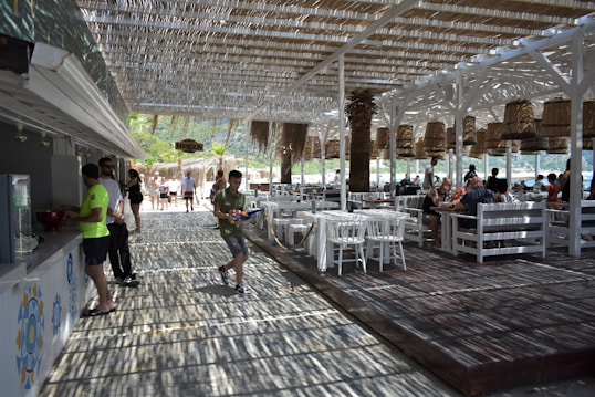 An outdoor dining area with white wooden furniture and a straw-covered roof allowing dappled sunlight through. People are seated at tables or walking around, and the area has a beachside ambiance with visible greenery and a glimpse of water in the background. Woven basket-style lights hang from the ceiling.
