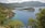 A picturesque coastal landscape featuring a serene bay surrounded by lush green hills and rocky cliffs. The water of the bay is a vibrant shade of blue, bordered by dense pine trees. A small island-like promontory extends into the water, creating a scenic and tranquil atmosphere.