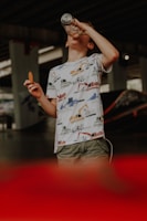 A young child is drinking from a clear water bottle while holding a cookie in the other hand. The child is wearing a white shirt with a colorful vehicle pattern and stands in an indoor setting with a shadowy background, possibly under a bridge or in a parking lot. The lower part of the image has a red blur, suggesting movement or an object close to the camera.