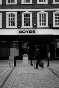 A black and white photo of a street scene featuring a shopfront named 'BOYES' with a person walking past it. The building has multiple large windows above the shop sign. The pavement is cobblestone, and there are signage boards near the store entrance.
