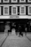 A black and white photo of a street scene featuring a shopfront named 'BOYES' with a person walking past it. The building has multiple large windows above the shop sign. The pavement is cobblestone, and there are signage boards near the store entrance.