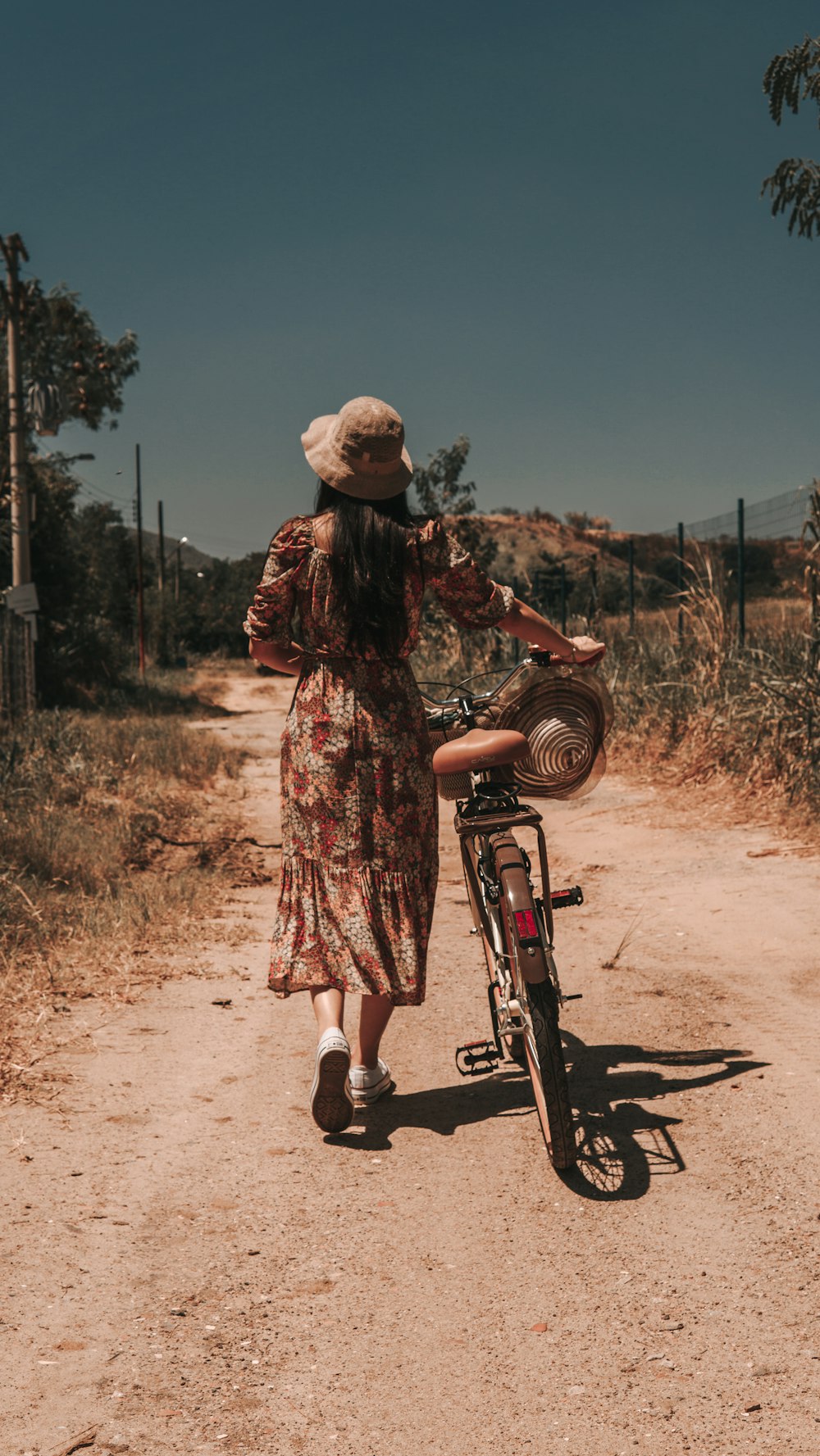 a woman in a dress and hat walking a bike down a dirt road