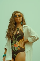 A woman with long curly hair wears orange sunglasses and a floral one-piece swimsuit. She is posing confidently with one hand on her hip, wearing a loose white shirt over her swimsuit. Her accessories include a turquoise beaded bracelet and earrings.