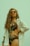 A woman with long curly hair wears orange sunglasses and a floral one-piece swimsuit. She is posing confidently with one hand on her hip, wearing a loose white shirt over her swimsuit. Her accessories include a turquoise beaded bracelet and earrings.