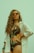A woman with long curly hair wears orange sunglasses and a floral one-piece swimsuit. She is posing confidently with one hand on her hip, wearing a loose white shirt over her swimsuit. Her accessories include a turquoise beaded bracelet and earrings.