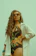 A woman with long curly hair wears orange sunglasses and a floral one-piece swimsuit. She is posing confidently with one hand on her hip, wearing a loose white shirt over her swimsuit. Her accessories include a turquoise beaded bracelet and earrings.