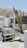 An outdoor café area features rustic wooden tables and benches under a light wooden pergola. A large cactus in a white pot is prominently placed in the foreground. Woven wicker light fixtures hang from the pergola roof, with high stools placed at a nearby bar.