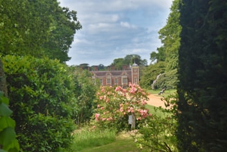 A large brick country house is situated amidst lush greenery and gardens. The foreground features vibrant pink flowers and a path leading towards the house. Tall trees frame the scene, creating a serene and picturesque setting.