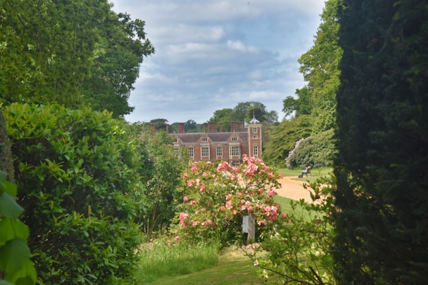 A large brick country house is situated amidst lush greenery and gardens. The foreground features vibrant pink flowers and a path leading towards the house. Tall trees frame the scene, creating a serene and picturesque setting.